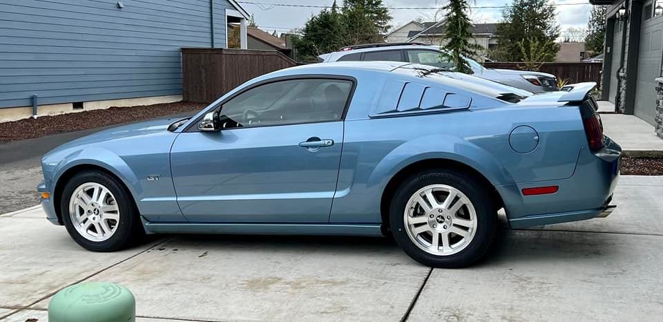 A blue Ford Mustang GT parked on a concrete driveway outside a house with blue siding.