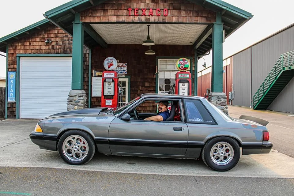 A man with a beard smiling and sitting in a gray vintage sports car, parked in front of a nostalgic Texaco gas station with two vintage gas pumps and a small office building.