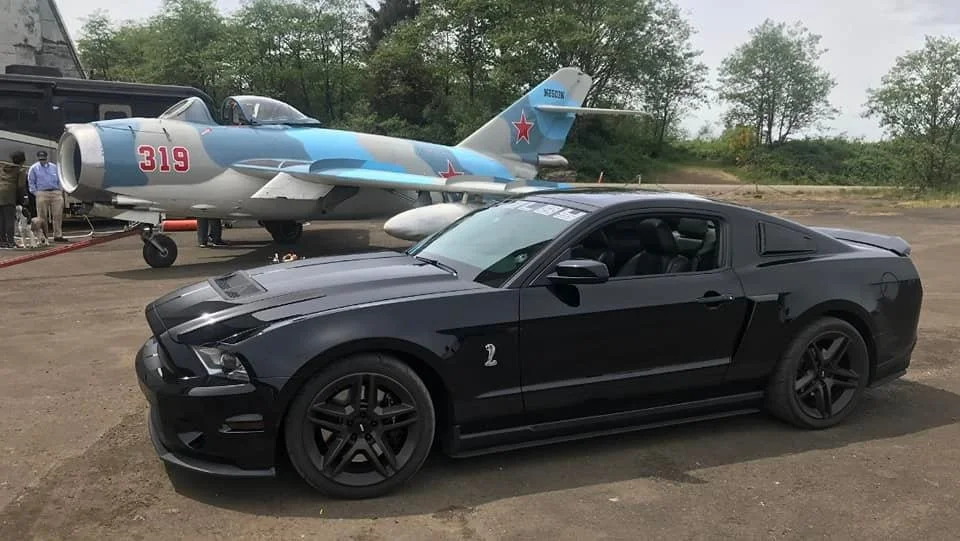A black Ford Mustang car parked outdoors with a gray and white fighter jet aircraft in the background. The jet has red star insignias and the number 319 on its nose.
