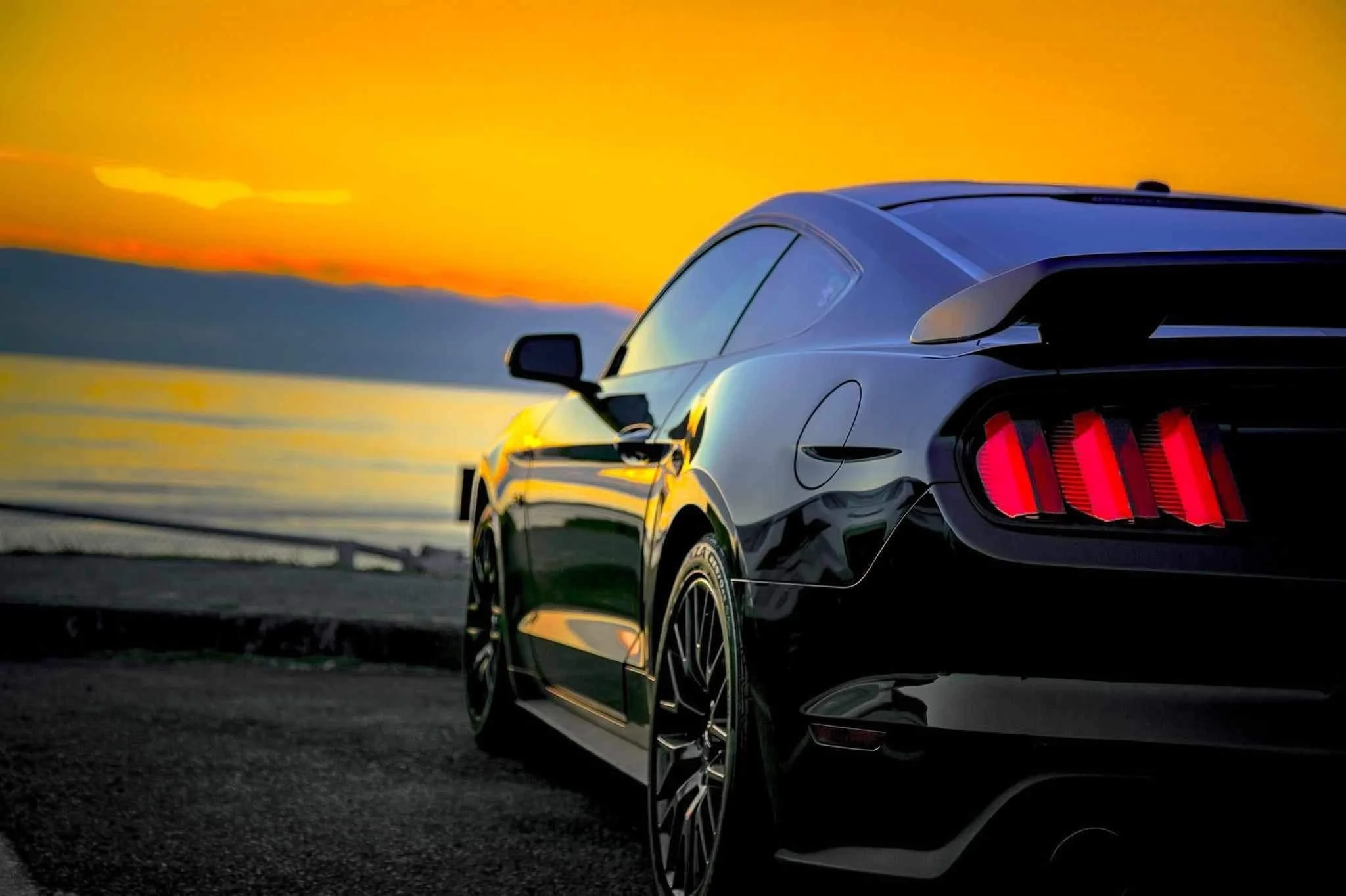 A black sports car parked on a dark surface near a body of water at sunset, with mountains in the background and a vibrant orange sky.