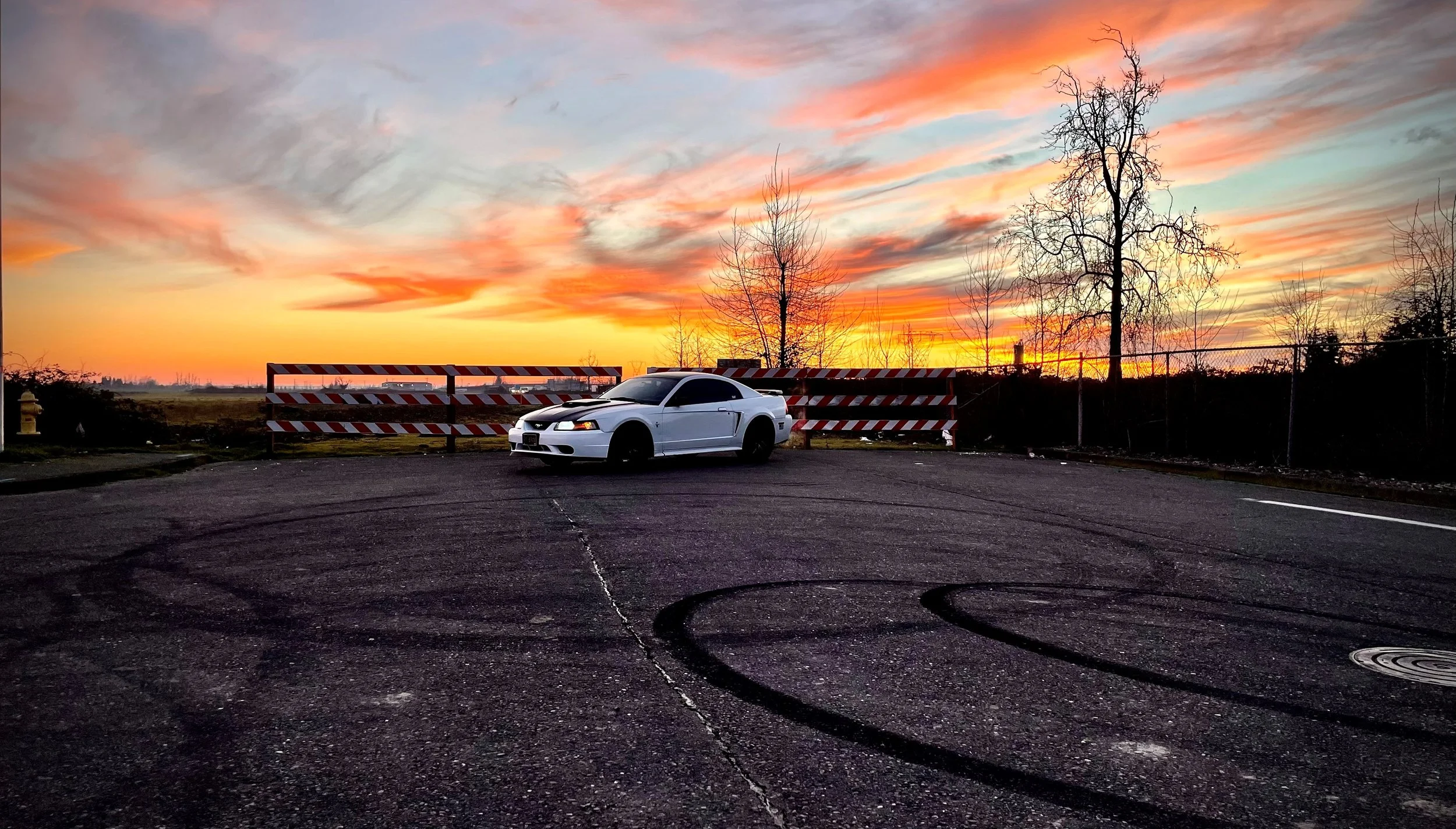 A white car parked in front of a barricade during a colorful sunset sky with clouds, leafless trees, and a chain-link fence in the background.