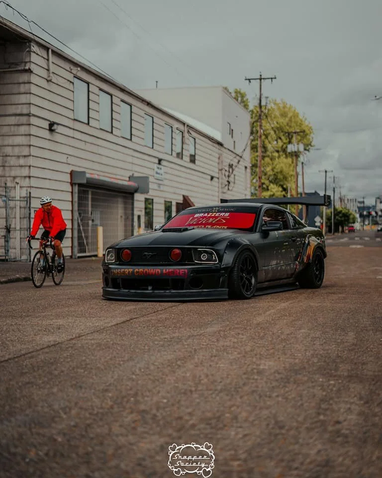 A black sports car with red tinted windows and a large rear wing is parked on a street. A cyclist in a red jacket and white helmet is riding past the car. The background features an industrial building with a garage door, trees, and utility poles und