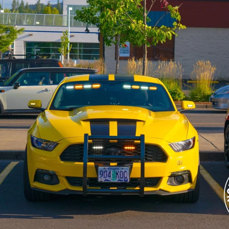 Yellow car with black racing stripes parked in a parking lot, with a bike rack attached to the front, Oregon license plate, and other vehicles in the background.