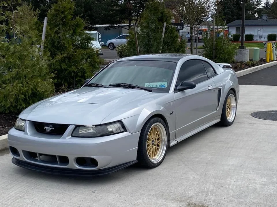 A silver Ford Mustang with gold wheels parked on a concrete driveway with green bushes and trees in the background.