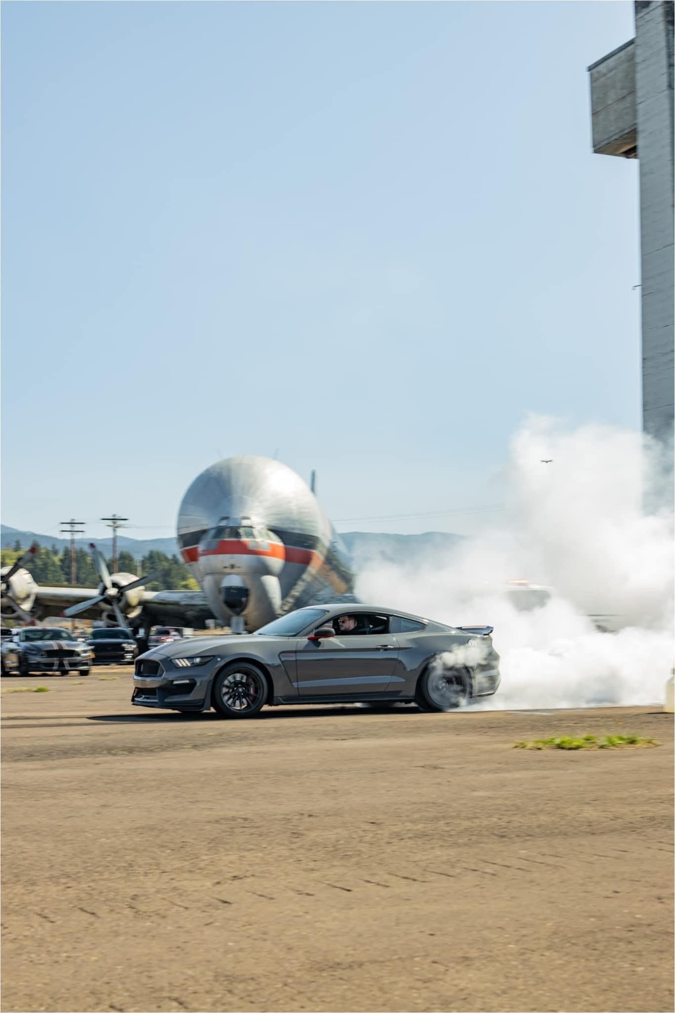 A black sports car driving at high speed with smoke coming from its tires on a paved lot, with a background of planes, a large missile, and a building.