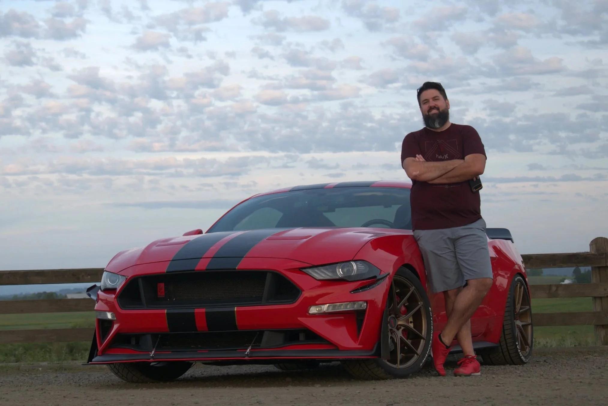 A man with a beard and mustache, wearing a maroon T-shirt and gray shorts, stands with arms crossed next to a red sports car with black racing stripes on a grassy area near a wooden fence, under a cloudy sky.