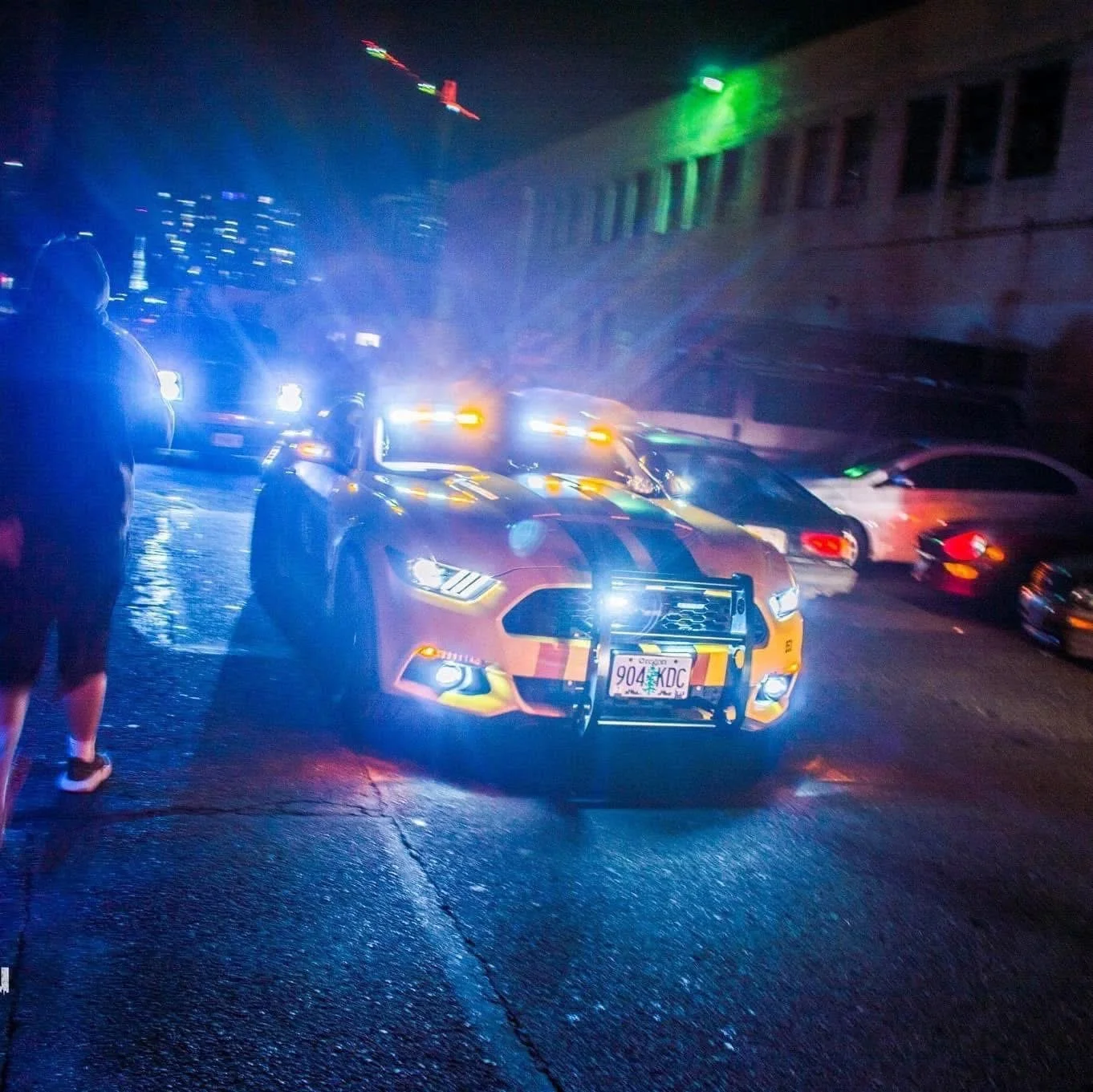 Night scene showing a police car with flashing lights parked on a wet street, with other cars and buildings in the background.