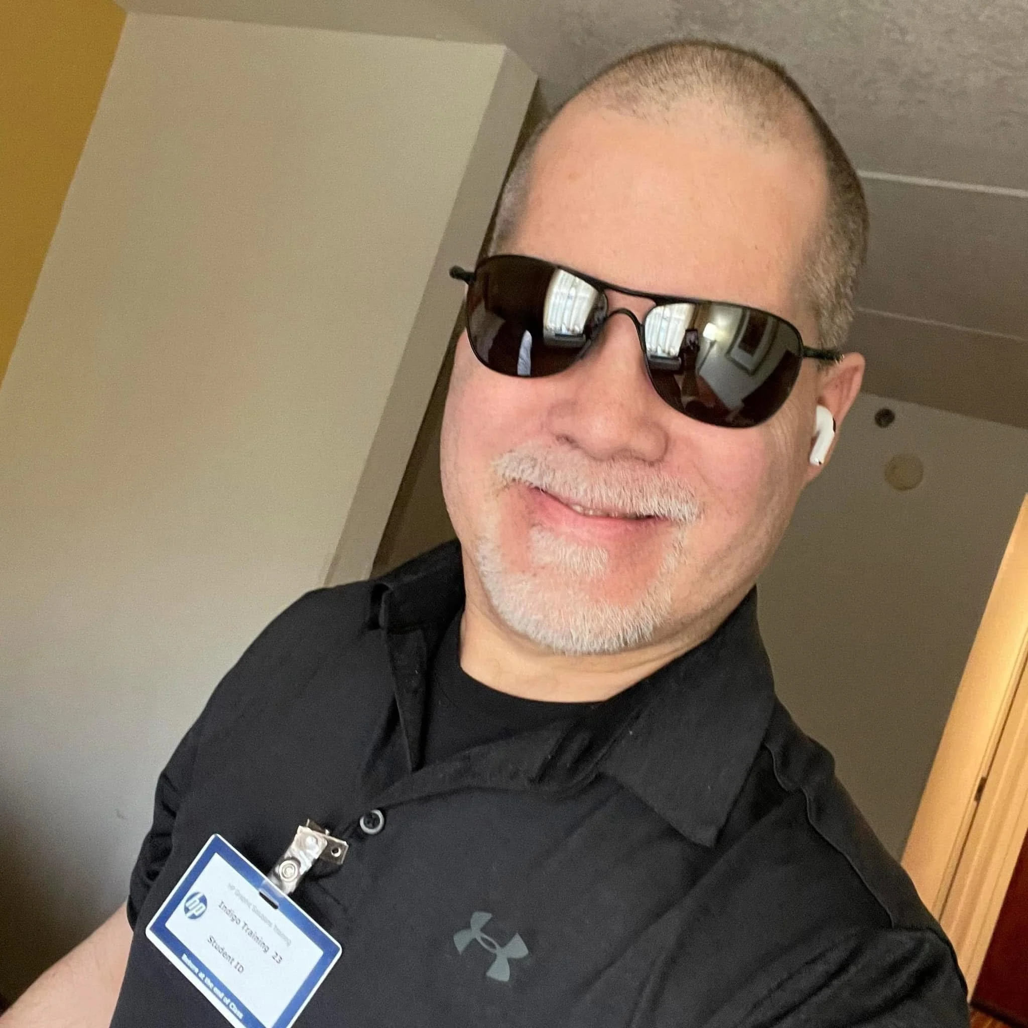 A man with a shaved head wearing sunglasses and a black shirt, with a name badge clipped to his shirt, standing indoors.