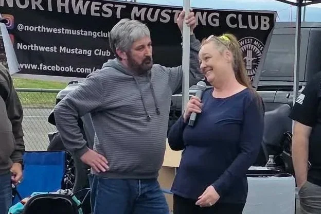 A woman holding a microphone speaking at an outdoor event, with a man standing next to her. In the background, there is a banner for the Northwest Mustang Club and a black canopy tent.