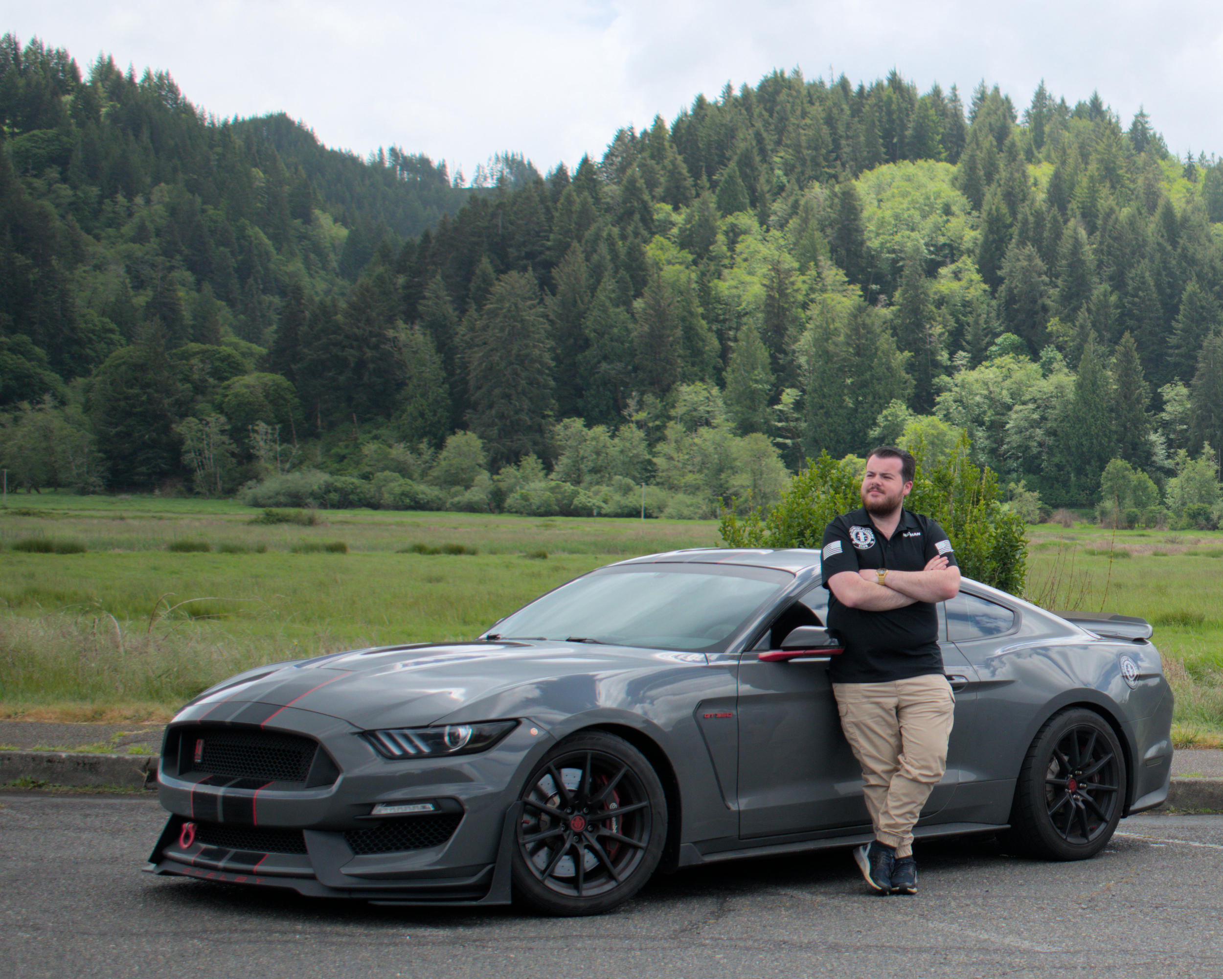 A man in a black shirt with a police patch on the sleeve standing with crossed arms next to a gray sports car, in a parking lot with greenery and a forested hillside in the background.