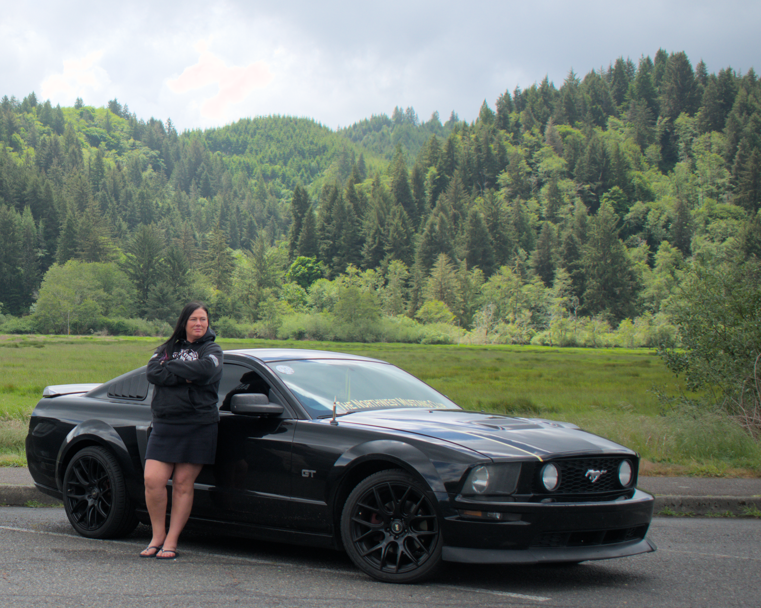 A woman with dark hair wearing a black jacket and shorts, standing in front of a black Ford Mustang GT car, with a forested mountain landscape and cloudy sky in the background.