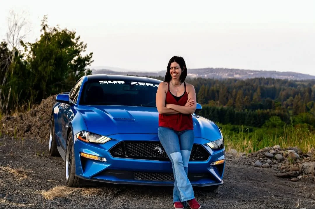 A woman with black hair in a red tank top and jeans standing in front of a blue sports car outdoors.