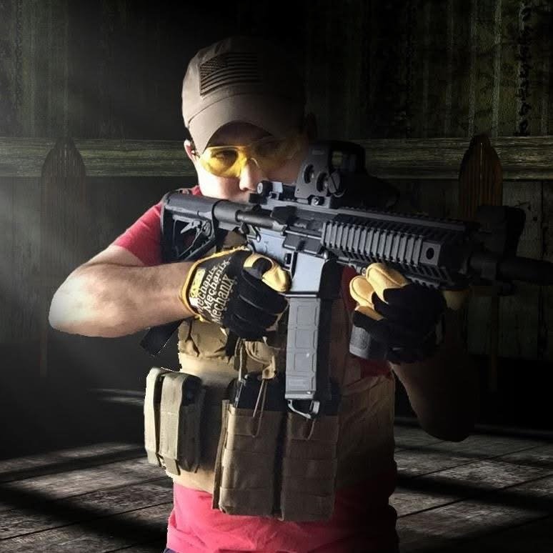 A person wearing a cap, yellow safety glasses, and camouflage gloves aiming a rifle in a wooden shooting range.