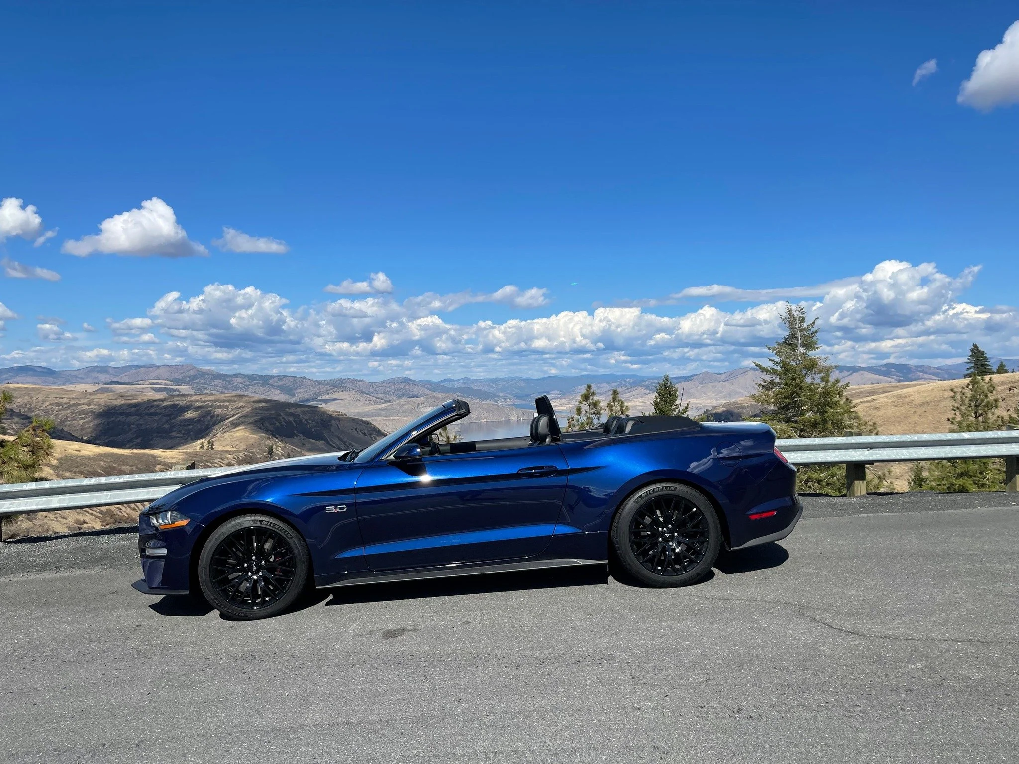 A blue convertible sports car parked on a mountain road with a guardrail, overlooking a valley with hills and scattered trees under a partly cloudy sky.