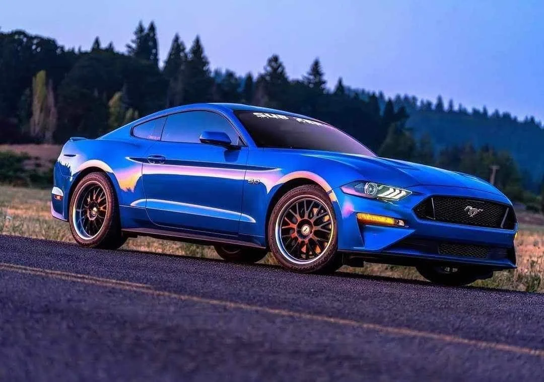 A blue sports car parked on a road with trees and mountains in the background during dusk.