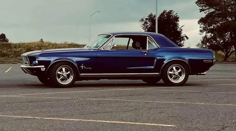 A vintage blue Ford Mustang parked in an empty parking lot with trees and a cloudy sky in the background.