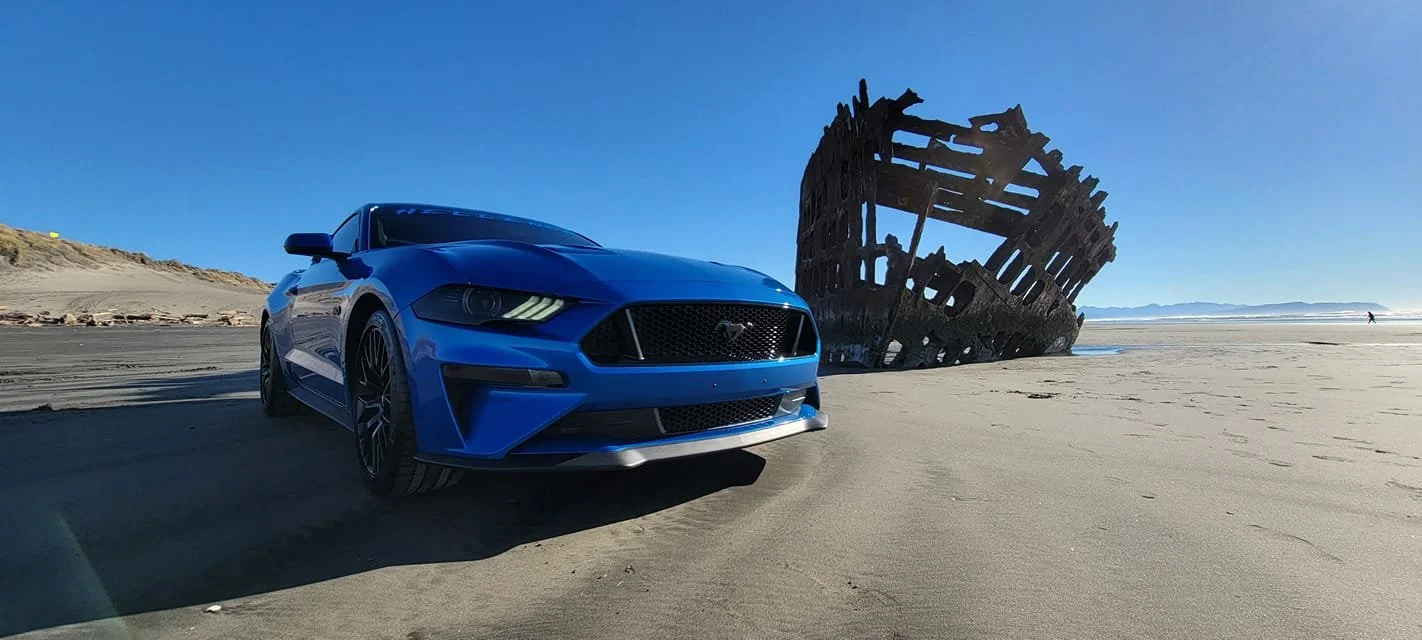 A blue sports car parked on a sandy beach with a rusted shipwreck in the background.