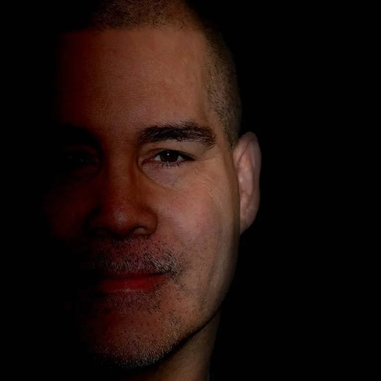 Close-up of a man's face, partially shadowed with a slight smile, short hair, and a thin beard.