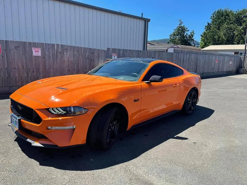 Orange Ford Mustang parked in an outdoor lot with a wooden fence and industrial buildings in the background.