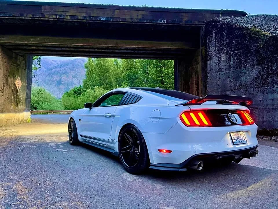 A white Ford Mustang car with black wheels parked under a bridge on a rural road with trees and mountains in the background.