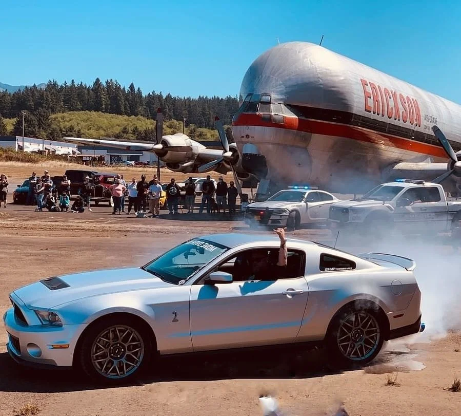A silver Ford Mustang Shelby GT is performing a burnout on a dirt airstrip, with smoke billowing from the tires. In the background, a large rocket with the words 'EPIC SOU' written on it is mounted on a transport vehicle, and a crowd of people is wat