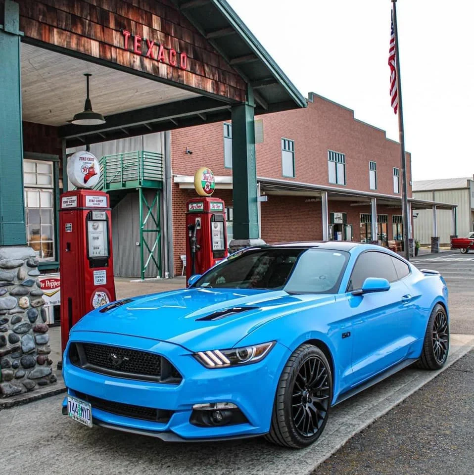 Blue Ford Mustang parked outside a Texaco gas station with vintage gas pumps, a brick building, and an American flag.