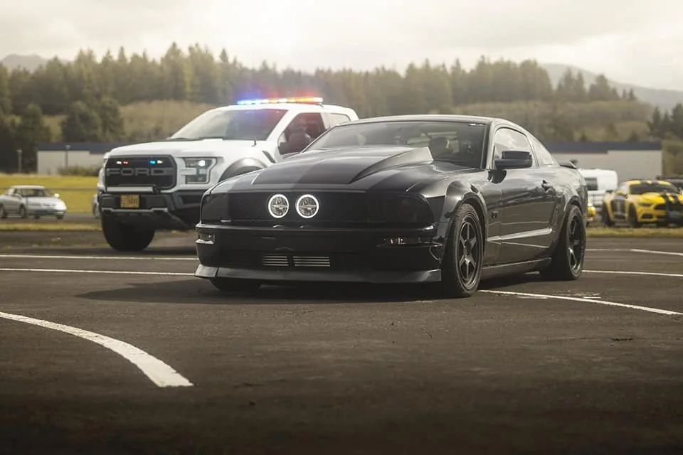 Black sports car parked in a lot with a police SUV with flashing lights in the background.