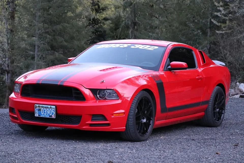Red Ford Mustang with black racing stripes, black wheels, and a license plate, parked on a gravel surface with trees in the background.