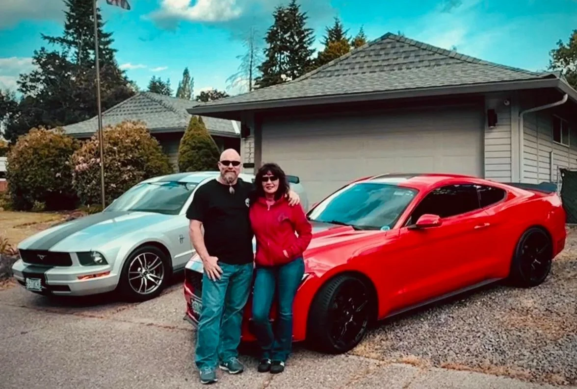 A man and woman standing in front of two cars, one silver and one red, in a driveway with a house in the background.