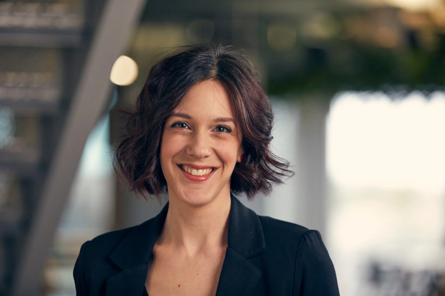 A woman with shoulder-length dark wavy hair smiling, wearing a black blazer, standing outside with blurred background of trees and building.