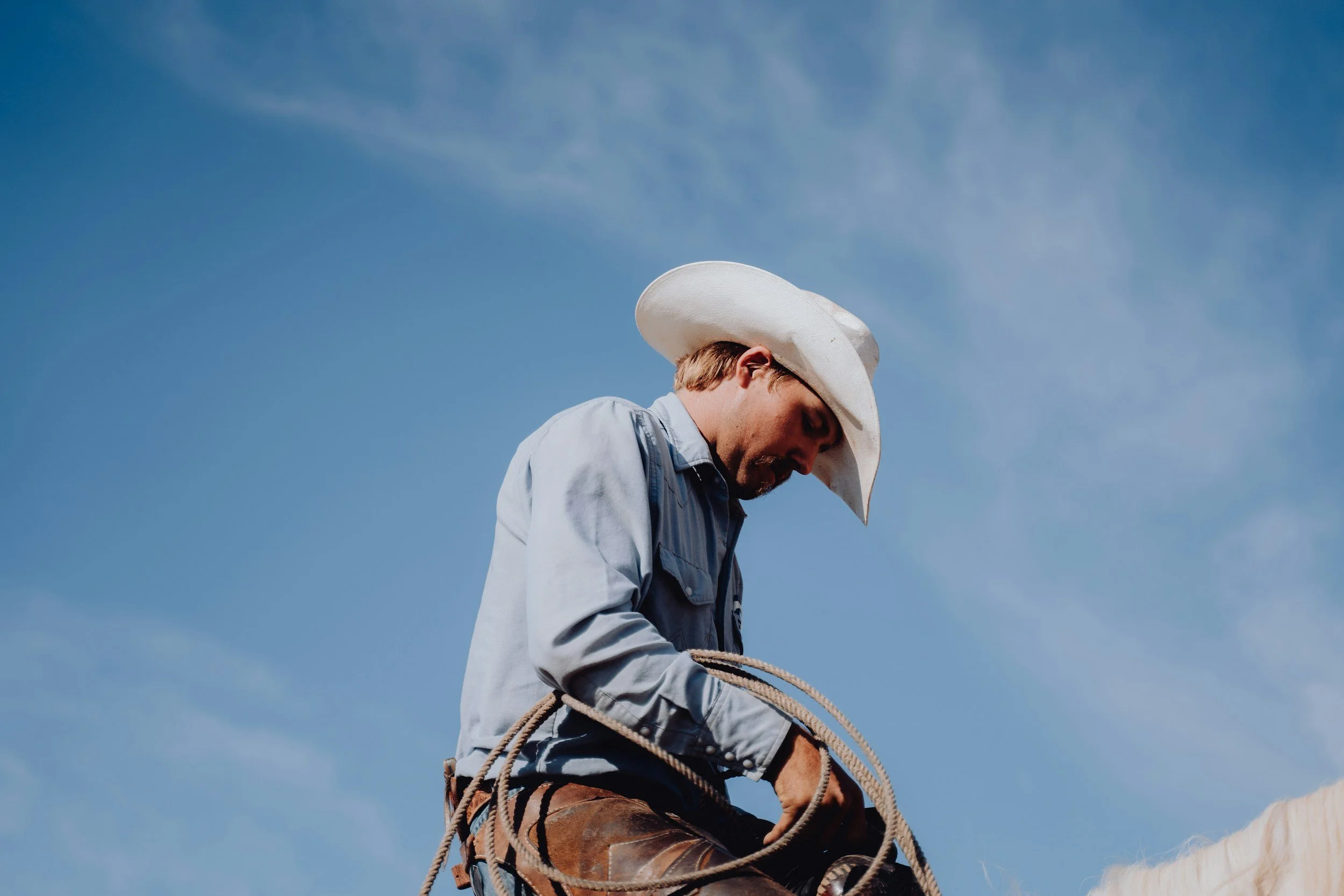 A man wearing a cowboy hat and denim shirt, holding a lasso, against a bright blue sky.