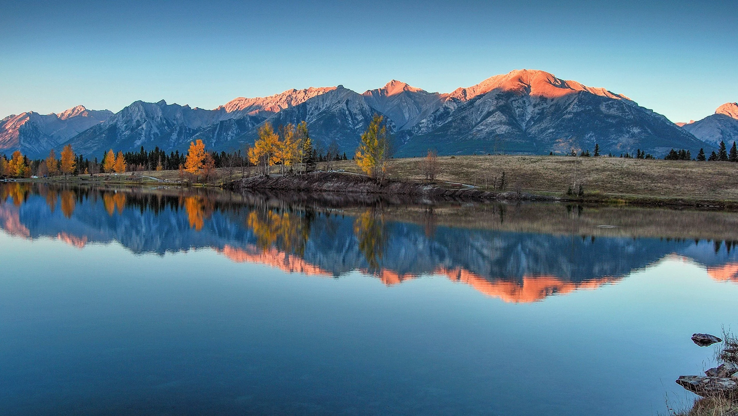 Snow-capped mountains reflected in a calm lake with yellow and green trees along the shoreline at sunset or sunrise.
