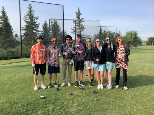 Eight women standing together on a golf course, holding golf clubs, with trees and a clear sky in the background.