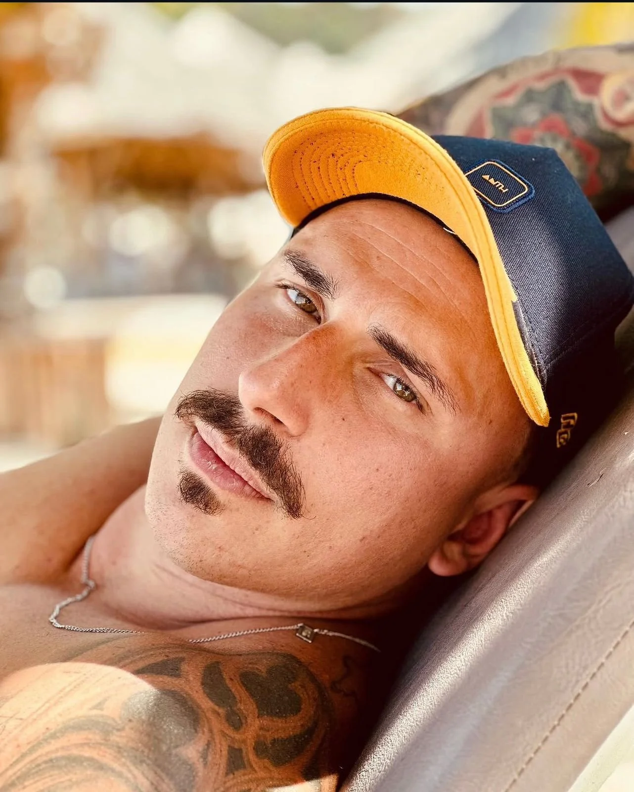 A young man with a mustache and goatee lying down, wearing a yellow and navy cap, with a tattooed arm showing, and a silver chain necklace, relaxing outdoors.