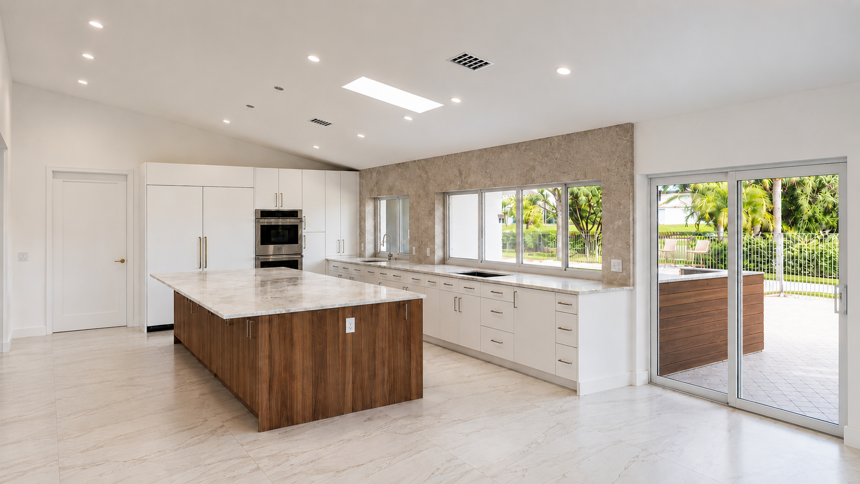 Modern kitchen with white cabinets, marble countertops, and a wooden island, large windows showing greenery outside, and a sliding glass door leading to an outdoor patio.