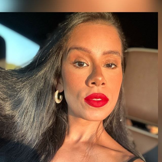 A woman with long dark hair, wearing gold hoop earrings and bright red lipstick, sitting in a vehicle with sunlight on her face.