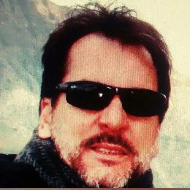 A man with dark hair and a beard, wearing sunglasses and a dark scarf, outdoors with a cloudy sky in the background.