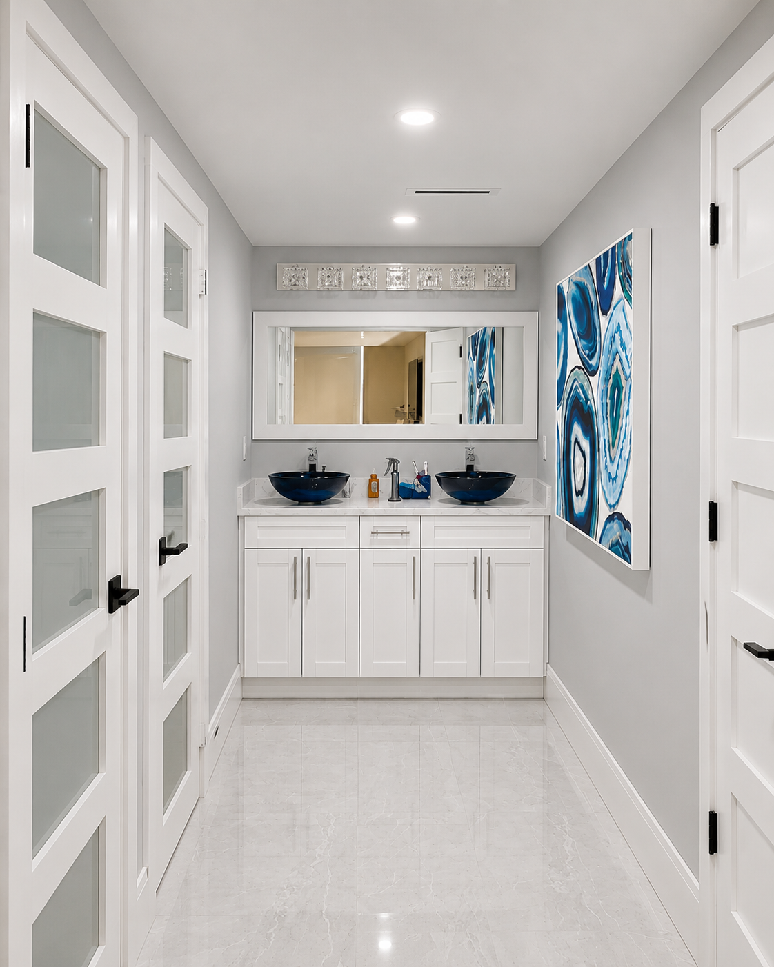 A modern bathroom with white cabinetry, two navy glass vessel sinks, a large framed mirror, abstract blue and white artwork on the wall, and frosted glass doors.