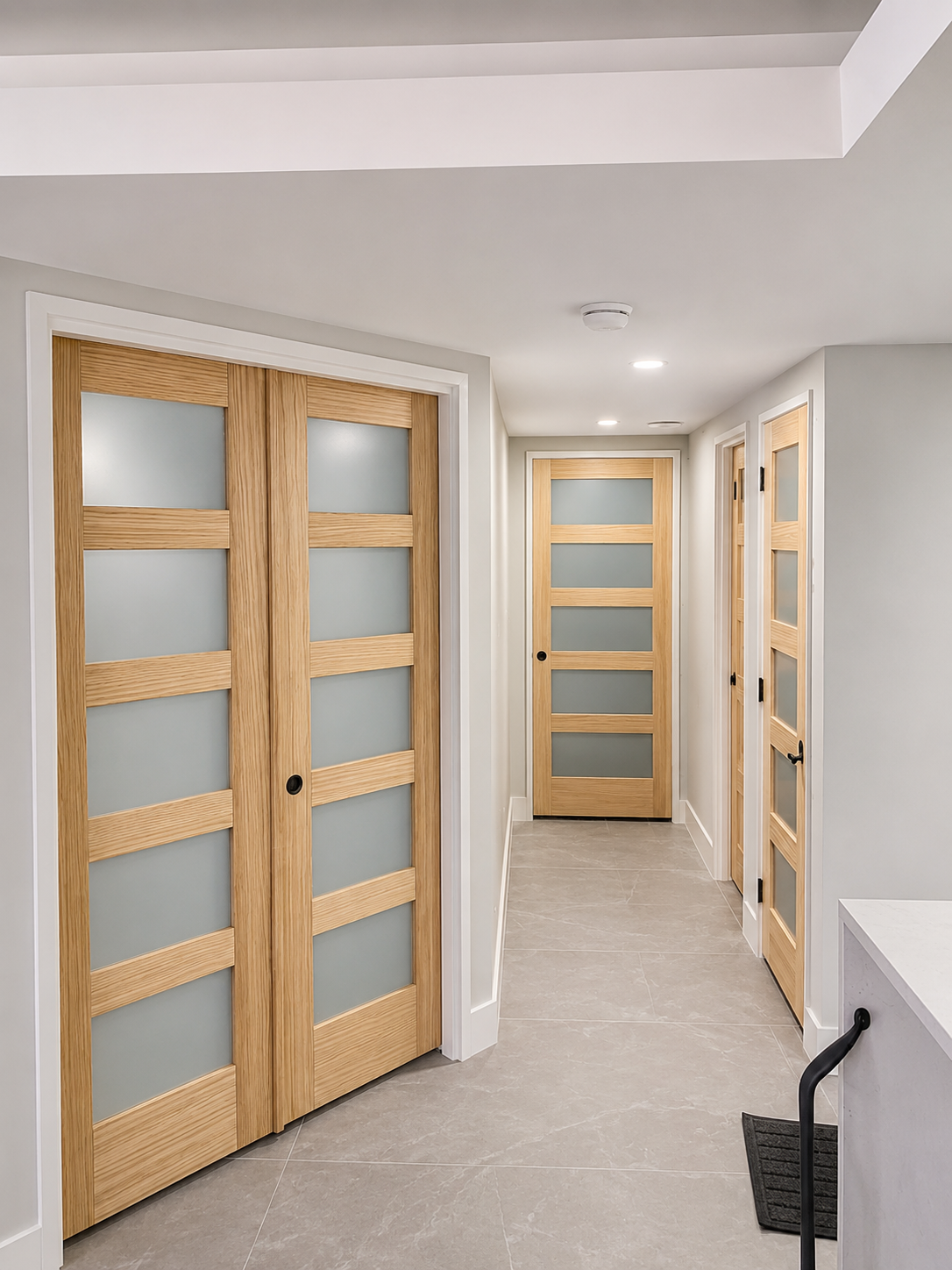 Hallway with three wooden doors with frosted glass panels and black hardware, neutral gray walls, beige tile flooring, and recessed lighting.
