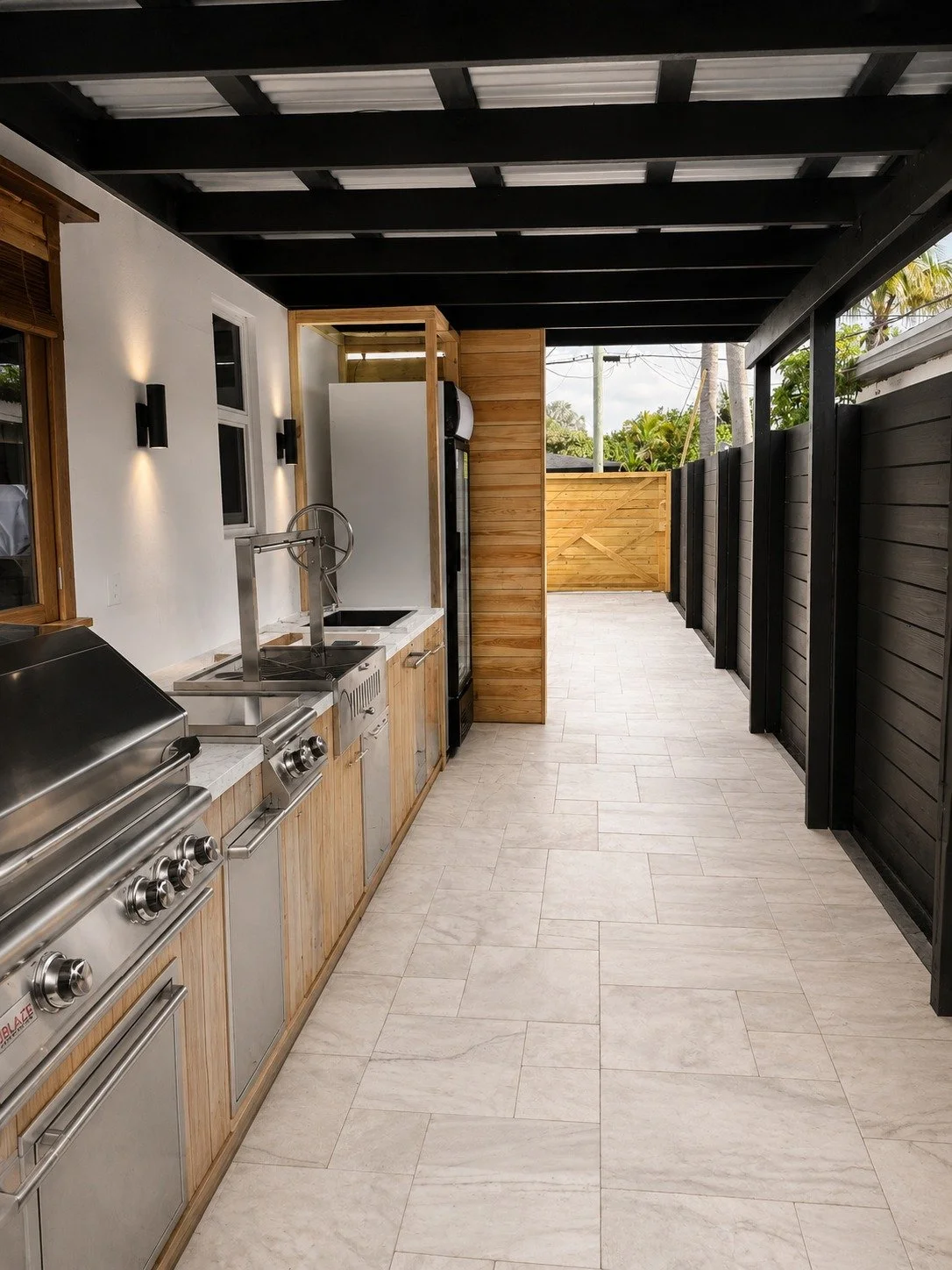 Outdoor kitchen with stainless steel grill, countertop, sink, and wooden cabinets, enclosed by black wooden fence and partial wooden wall, with a covered roof and tropical trees visible beyond.