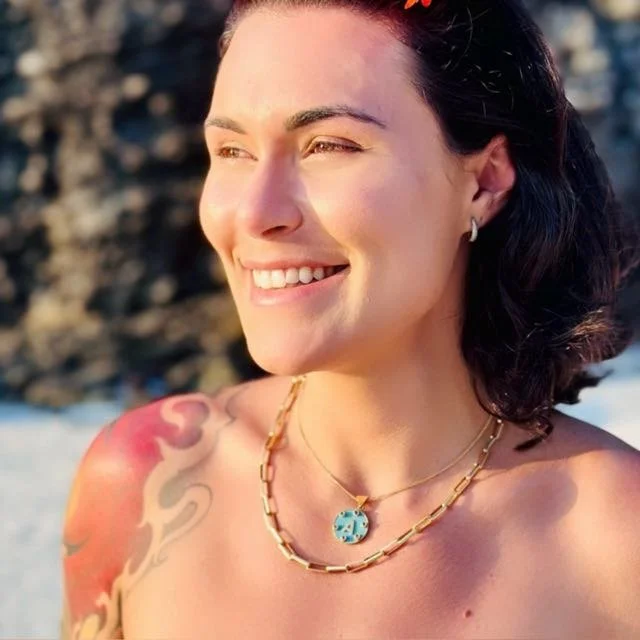 A smiling woman with dark hair, wearing jewelry, and a colorful tattoo on her shoulder, outdoors near a rocky shoreline