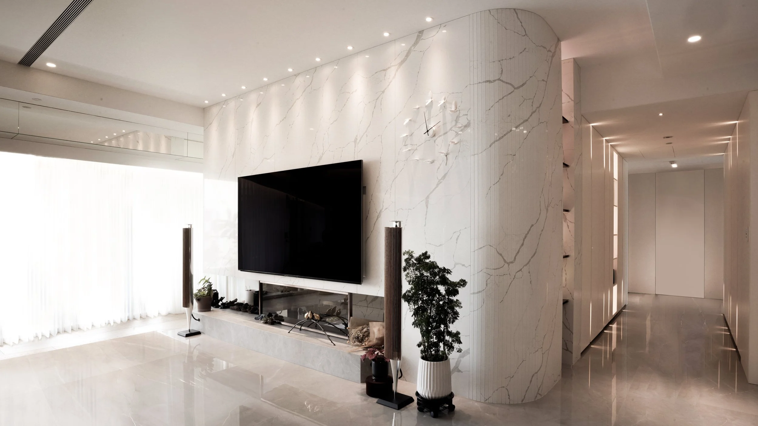 Modern living room with a large black flat-screen television mounted on a white marble wall with gold veining, flanked by tall speakers and potted plants. The room features a glossy white floor, built-in storage shelves along the curved wall, and a hallway with recessed lighting.