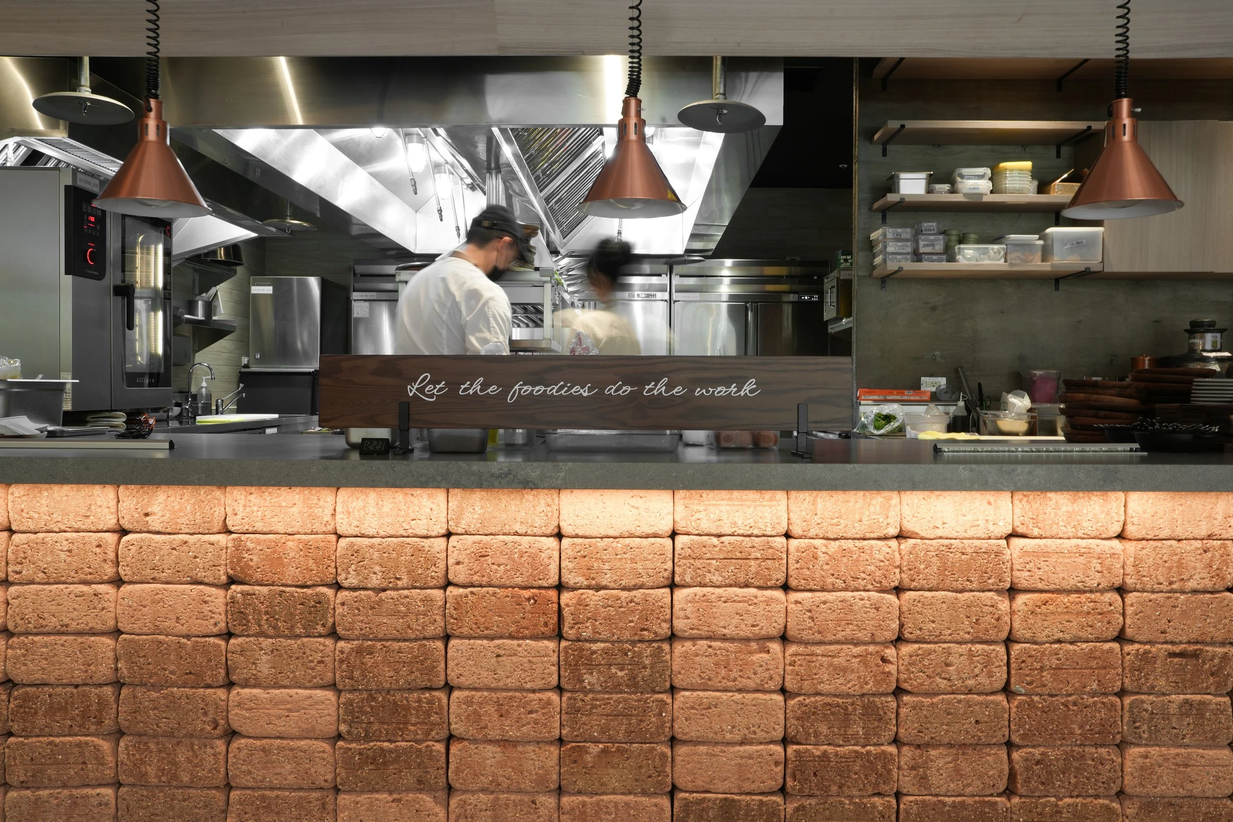 A commercial kitchen with two chefs working behind a counter. The front of the counter is made of stacked bricks, and a sign reads "Let the foodies do the work." Shelves with various containers and kitchen tools are on the right side.