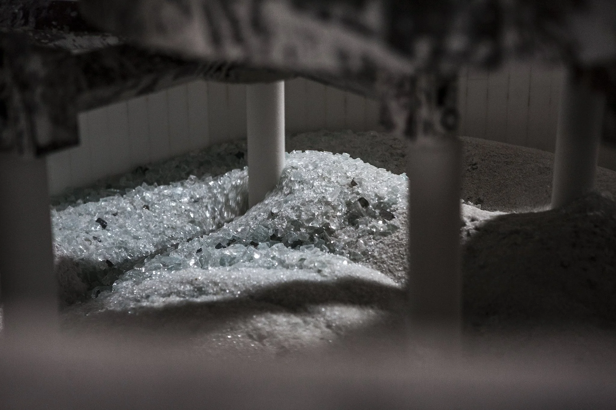 Crushed glass bottles and shards beneath a cardboard box, supported by white metal bars, with sand on one side.