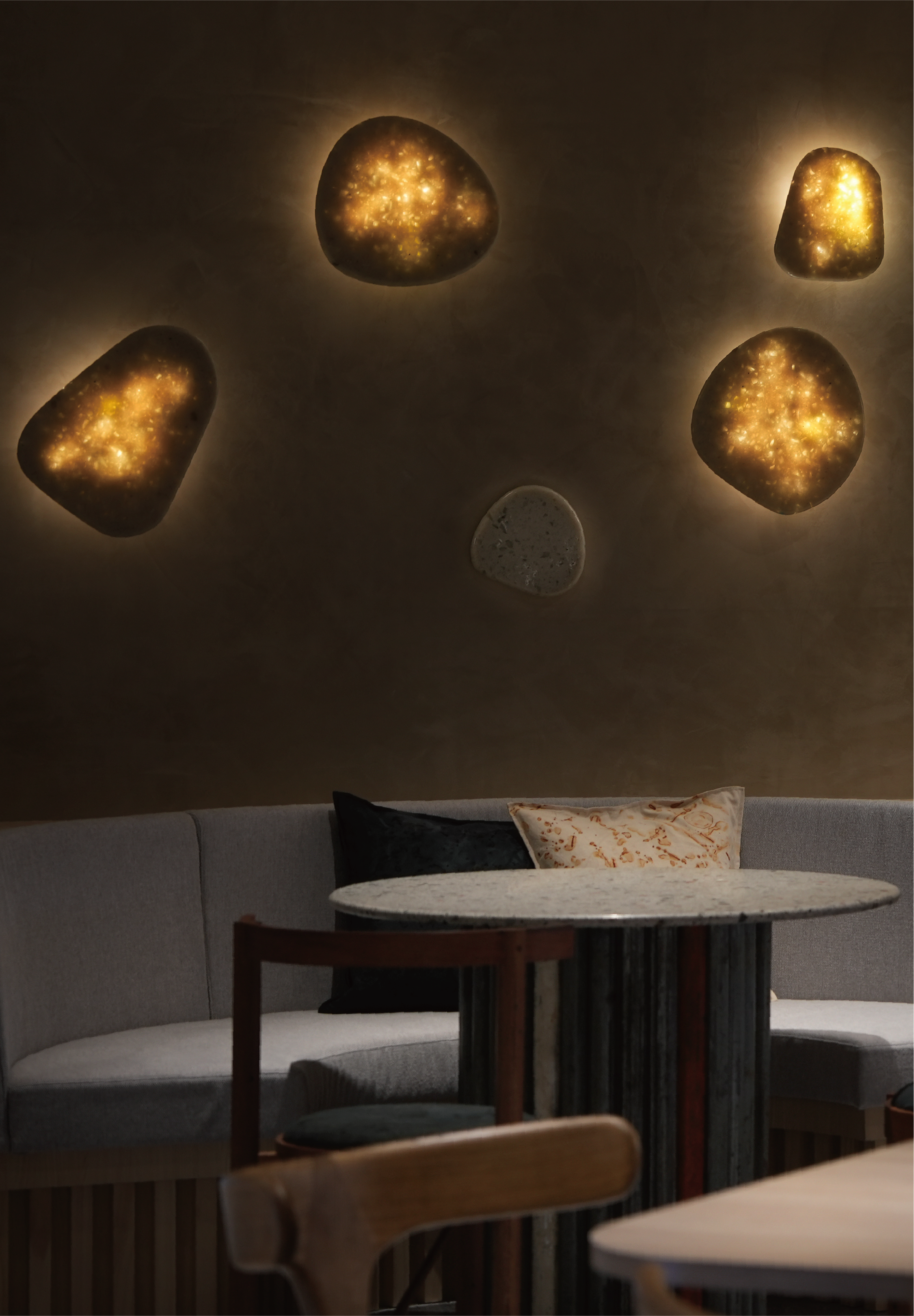 A dimly lit interior with a curved gray booth, a circular table with a black base, and a decorative wall with illuminated stones resembling stars or planets.