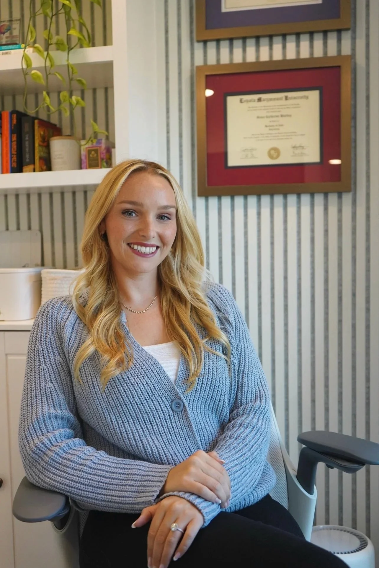 A smiling young woman with long blonde hair, wearing a gray cardigan and a white shirt, sitting in an office chair in front of bookshelves and framed certificate on the wall.
