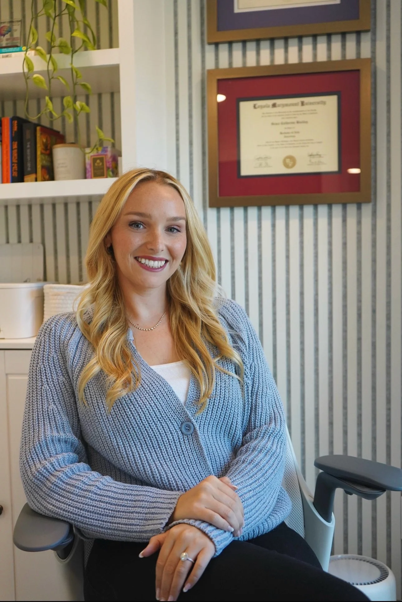 Smiling woman with long blonde hair sits in an office chair, diploma on wall behind her, books and plant on shelves to the left.