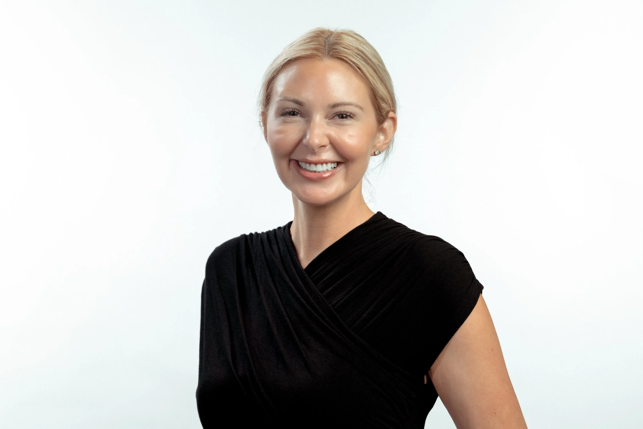 A woman with blonde hair styled back, smiling, wearing a black dress, against a plain white background.