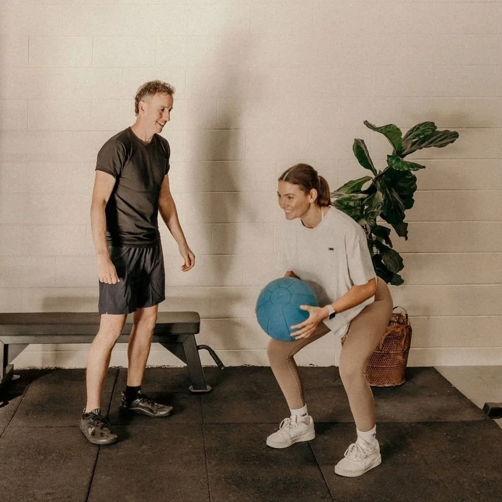 A woman in white shirt and beige pants doing squats with a blue exercise ball while a man in black workout clothes watches her in a gym with a black mat and a large green plant in the background.