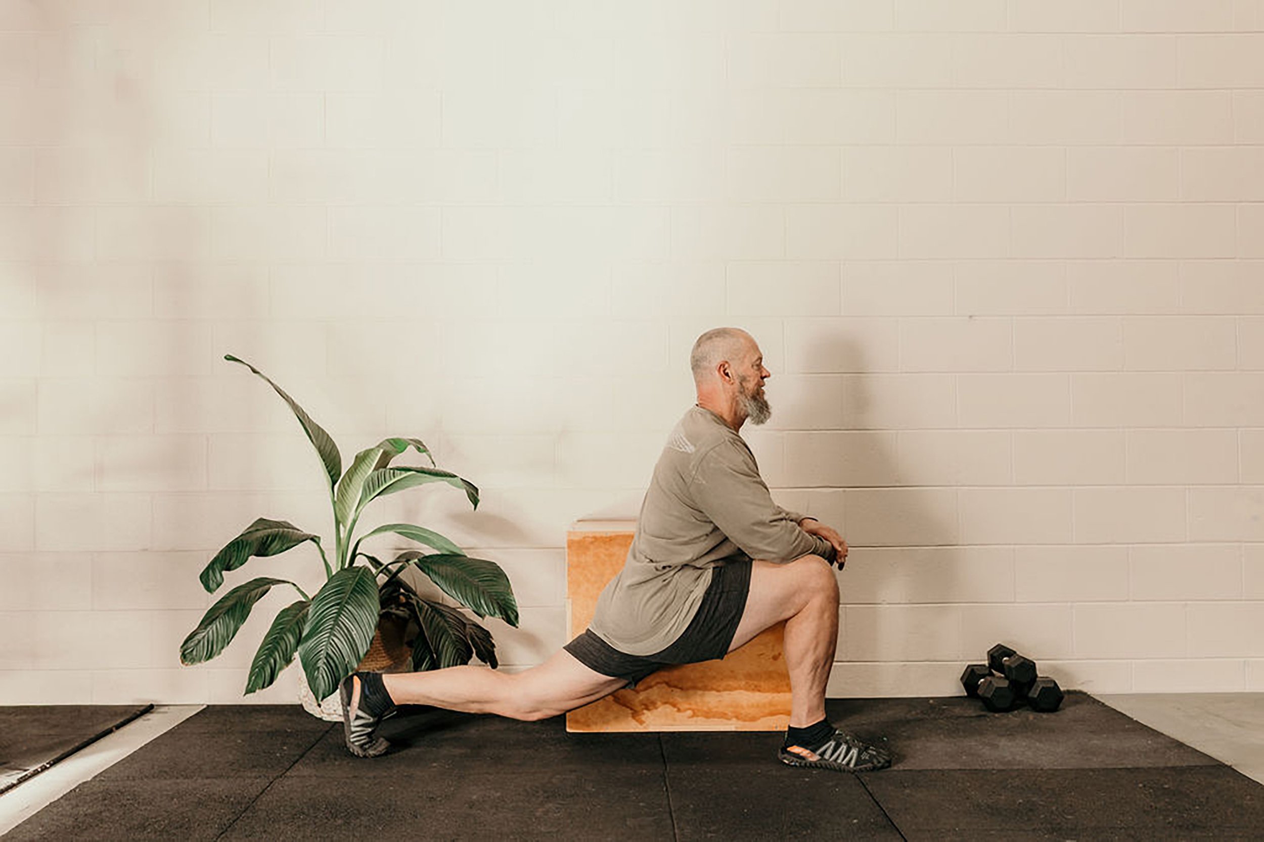 A man with a beard doing a yoga pose in a gym, with a plant and some dumbbells nearby. 4b Gym Pambula NSW.